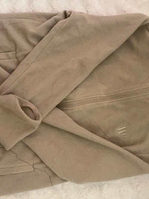 Ridtrlja - Mysig beige ridtröja i mjuk fleece med skärp i midjan och bred krage. Perfekt för att chilla hemma och hålla sig varm under kalla morgnar. Enkel och stilren design utan synliga mönster eller detaljer.