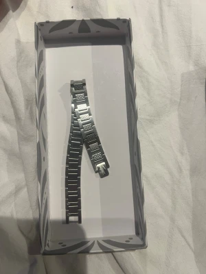 Lacoste Silverfärgad länkarmband  - Snyggt silverfärgat länkarmband i metall. Armbandet har en klassisk design med rektangulära länkar och detaljerade mönster på vissa länkar. Perfekt för dig som vill byta ut eller uppgradera din klocka med en stilren metallänk. Ingen box