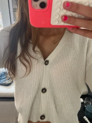 Vit ribbstickad kofta med knappar - Vit ribbstickad kofta med v-ringning och stora mörka knappar framtill. Koftan har långa ärmar och en avslappnad passform, perfekt att slänga på sig över en t-shirt. Materialet ser mjukt och bekvämt ut.