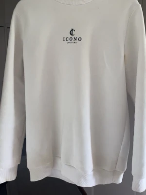 Vit sweatshirt från Icono Couture - Vit sweatshirt med rund hals från Icono Couture. Framsidan har en liten logga på bröstet och baksidan pryds av ett stort, detaljerat svart tryck med mönster och texten 'Atelier Icono'. Tröjan har ribbade muddar vid ärmslut och nederkant.