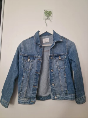 Blå jeansjacka från Zara - Klassisk blå jeansjacka från Zara i storlek 140. Jackan har en tidlös design med skjortkrage, knappstängning framtill och detaljerade sömmar. Perfekt att slänga över en hoodie eller t-shirt för en avslappnad look.