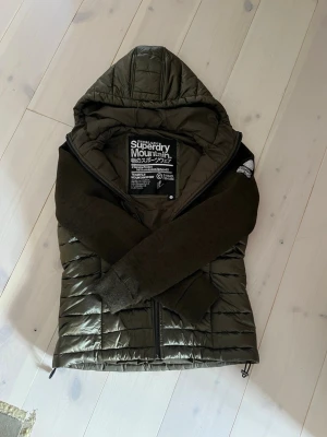 Superdry Jacka - Snygg olivgrön pufferjacka från Superdry med huva och dragkedja framtill. Jackan har quiltad kropp och mjuka, stickade ärmar i mörkare ton. Perfekt för lite kallare dagar och har en sportig, modern look. Står storlek M men sitter mer som en S.