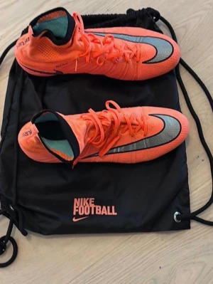 Nike Mercurial Superfly IV FG 641858-804   - Snygga Nike Mercurial fotbollsskor i en stark orange färg med svarta och silverfärgade detaljer. Skorna har hög ankelkrage i stickat material och klassisk snörning. Perfekta för dig som vill ha grym touch och kontroll på planen. Levereras med svart Nike Football-påse.NIKE - Nike Mercurial Superfly IV FG 641858-804  Elite stl 43