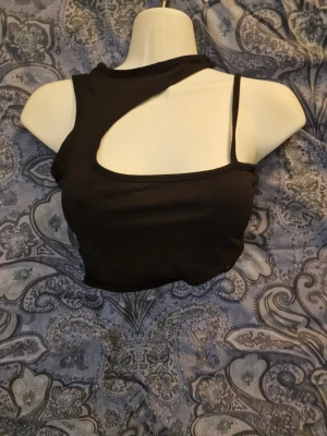 Svart asymmetrisk croppad topp - Säljer en svart croppad topp med asymmetrisk design och cut-out detalj vid axeln. Toppen har en axelbandslös sida och ett smalt axelband på andra sidan, vilket ger en unik och trendig look. Perfekt för dig som vill sticka ut med din stil.