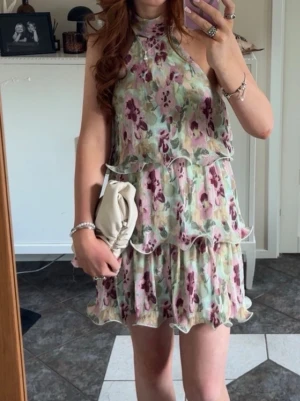 Blommig kortklänning med volang - Söt kortklänning med blommigt mönster i lila, rosa och grönt. Klänningen har hög hals, är ärmlös och har volangdetaljer längs kjolen. Perfekt för sommarens alla tillfällen och ger en romantisk vibe.