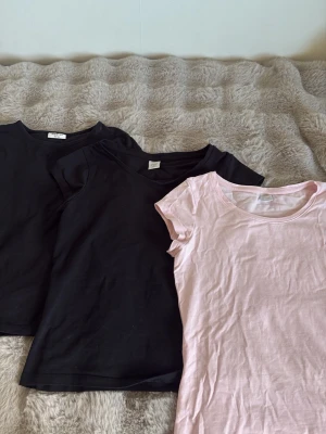 Två svarta och en ljusrosa t-shirt - Säljer tre basic t-shirts i mjuk bomull, två svarta och en ljusrosa. Alla har rund halsringning och korta ärmar. Perfekta att matcha till jeans eller kjol för en enkel och clean look.