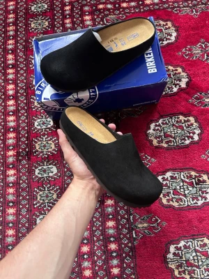 Svarta Birkenstock tofflor i mocka - Säljer ett par svarta tofflor från Birkenstock i mjuk mocka med klassisk slip-in design. De har en bekväm innersula och robust yttersula, perfekta för chill dagar hemma. Stilsäkra och tidlösa, passar till det mesta i garderoben.