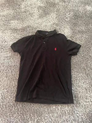 Svart Ralph Lauren piké - Säljer en svart Ralph Lauren pikétröja med klassisk krage och två knappar framtill. Tröjan har en liten röd broderad logga på bröstet och är tillverkad i mjuk bomull. Passformen är normal och modellen är kortärmad, perfekt till jeans eller shorts.