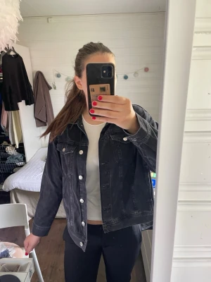 Svart jeansjacka - Säljer en urtvättad svart jeansjacka med klassisk krage och silverfärgade knappar. Jackan har två bröstfickor med lock och knapp samt raka ärmar. Perfekt för en avslappnad och trendig look.