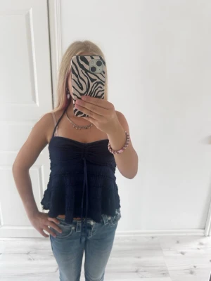Mörkblå volang topp med knyt - Söt mörkblå topp med volanger och knyt framtill. Toppen har tunna axelband och halterneck-knyt i nacken. Perfekt till jeans eller kjol för en trendig look. Materialet känns lätt och luftigt, perfekt för varma dagar.