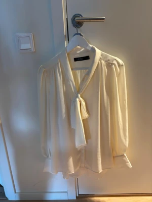 Krämvit blus med knyt från Ralph Lauren - Stilren krämvit blus från Ralph Lauren med knytband i halsen och långa, lätt puffade ärmar. Blusen har en mjuk och är tillverkad i ett  material som ger en lyxig känsla.
