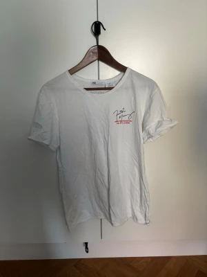 Vit t-shirt från Zara med tryck - Vit t-shirt från Zara med ett litet tryck och signatur på bröstet. Klassisk rund hals och korta ärmar. Tillverkad i mjuk bomull som känns skön mot huden. Perfekt för en avslappnad och stilren look.