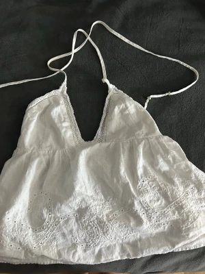 Vit sommartopp med spets - Söt vit linnetopp med tunna axelband och v-ringning. Toppen har fina spetsdetaljer längs urringningen och broderade mönster nertill. Tillverkad i ett lätt och luftigt material, perfekt för varma dagar.