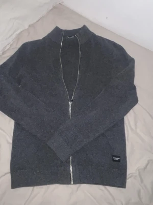 Mörkgrå ziptröja från Jack & Jones - Passar även storlek S.                    Snygg mörkgrå ziptröja från Jack & Jones med hög krage och hel dragkedja framtill. Tröjan har ribbade muddar vid ärmslut och nederkant samt en diskret logolapp nertill. Perfekt för lager på lager och enkel att matcha.