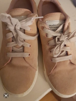 Beiga sneakers från Marc Jacobs - Snygga beiga sneakers från Marc Jacobs med vit sula och vita skosnören. Ovandelen är i mockaliknande material och har en stilren design med diskret logga på plösen. Perfekta för en trendig och avslappnad look.