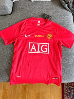Manchester United Ronaldo t-shirt röd - Säljer en röd Manchester United 2008 UCL final t-shirt med tryck 'AIG' på bröstet och 'Ronaldo 7' på ryggen. Tröjan har korta ärmar, rund hals och är tillverkad i ett lätt och sportigt material från Nike. Perfekt för fotbollsfans! Passar för folk som har storlek M 
