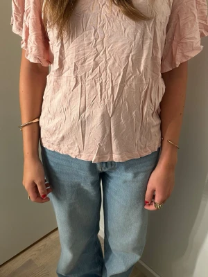 Ljusrosa topp med volangärmar - Söt ljusrosa topp med korta volangärmar och avslappnad passform. Tillverkad i mjukt material som känns skönt mot huden. Perfekt att styla med jeans eller kjol för en trendig look.