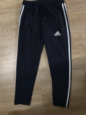 Svarta Adidas mjukisbyxor med vita ränder - Svarta mjukisbyxor från Adidas med klassiska vita ränder längs sidorna och Adidas-logga på låret. Byxorna har resår i midjan med dragsko och två sidofickor. Perfekta för en avslappnad och sportig stil.