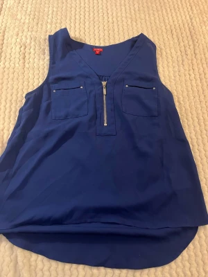 Blå ärmlös topp från Guess - Säljer en snygg blå ärmlös topp från Guess med v-ringning och dragkedja framtill. Toppen har två bröstfickor med små silverfärgade detaljer och är tillverkad i ett lätt och mjukt material. Perfekt för varma dagar eller att styla lager på lager.