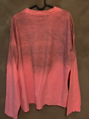 Rosa långärmad topp Acne Studios - Säljer en rosa långärmad topp från Acne Studios med texten 'Acne Studios STOCKHOLM 1996' på bröstet. Tröjan har en snygg ombre-effekt från mörkare till ljusare rosa och är tillverkad i mjuk bomull. Perfekt för dig som gillar streetwear vibes.