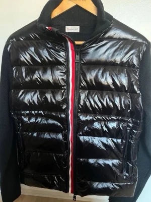 Svart dunjacka från Moncler - Snygg svart dunjacka från Moncler med glansig front och stickade ärmar. Jackan har dragkedja framtill med röd och vit detalj, två sidofickor med dragkedja och Moncler-logga på ärmen. Perfekt för kalla vinterdagar och riktigt trendig look.