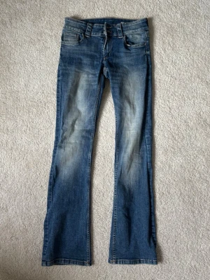 Blåa bootcut jeans med slitningar - Säljer ett par blåa bootcut jeans med snygga slitningar och tvättade detaljer. Jeansen har låg midja, klassiska fem fickor och stängs med dragkedja och knapp. 
