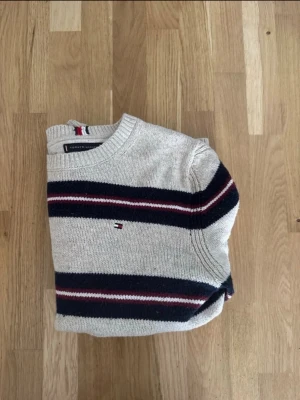 Randig stickad tröja Tommy Hilfiger - Snygg stickad tröja från Tommy Hilfiger i ljusgrått med marinblå, röda och vita ränder. Klassisk rund halsringning och diskret logga broderad på bröstet. Perfekt till jeans för en avslappnad men stilren look.