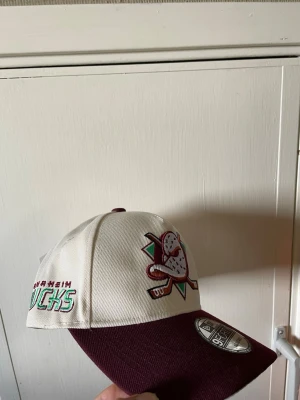 Anaheim Ducks keps NHL - Snygg keps från New Era med Anaheim Ducks-logga framtill. Kepsen har vit krona och vinröd skärm, samt broderade detaljer i grönt, lila och svart. Klassisk 9FIFTY snapback-modell.