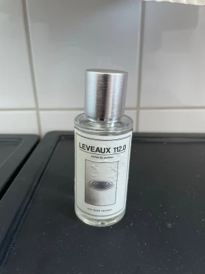 Leveaux 112.0 parfymflaska - Inspirerad av YSL Y Intense. Minimalistisk parfymflaska med transparent glas och silvrig metallkork. Etiketten är vit med svart text och en abstrakt bild. Flaskan har en stilren och modern design som passar perfekt på sminkbordet.