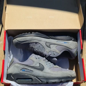 Nike Air Max 90 grå sneakers - Snygga Nike Air Max 90 sneakers i olika nyanser av grått med blå detaljer. Skorna har en klassisk låg siluett, mesh och mocka på ovandelen samt den ikoniska Air Max-dämpningen i sulan. Perfekta för dig som gillar streetwear och sportig stil.