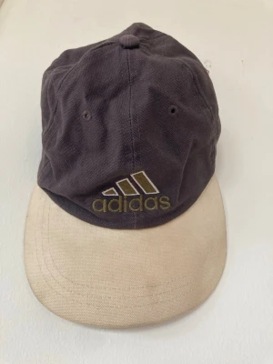 Vintage Adidas keps  - Snygg keps från Adidas i grått tyg med beige skärm. Broderad Adidas-logga framtill i guld och vitt. Justerbar rem baktill för perfekt passform. Tillverkad i 100% bomull för en skön känsla. Lite smutsig skärm, inte testat att tvätta bort. 