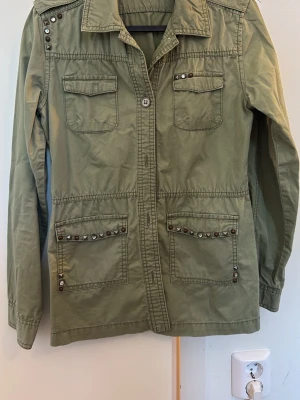 Olivgrön overshirt med nitar - Snygg olivgrön overshirt med fyra fickor framtill och coola nitar och metallknappar som detaljer på axlar och fickor. Rak passform, klassisk krage och långa ärmar. Tillverkad i ett slitstarkt bomullstyg som ger en avslappnad look. Storlek 158 barn.