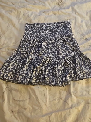 Blå blommig kjol  - Supersöt blå kjol från 157 med vitt blommönster. Kjolen har smock i midjan för en bekväm passform och volang nedtill som ger en flowy känsla. Perfekt för dig som gillar en feminin och avslappnad stil.