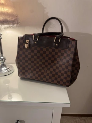 Brun rutig handväska med röda detaljer - Snygg handväska i brunt rutigt mönster med mörkbruna handtag och guldfärgade metalldetaljer. Väskan har en dragkedja upptill, röd insida och flera fack för smart förvaring. Perfekt accessoar för dig som gillar klassisk och trendig stil.