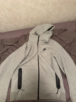Nike tech  - Snygg grå hoodie från Nike med svart dragkedja och två fickor med dragkedjor. Klassisk Nike-logga på bröstet och stor huva. Tillverkad i mjukt sweatshirtmaterial som är perfekt för chill eller träning. Enkel och stilren design som funkar till det mesta.