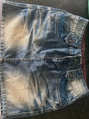 Kort jeanskjol med paljettficka - Säljer en kort jeanskjol i blått med slitningar och coola detaljer. Kjolen är aldrig använd och den är i storlek XL men är liten i storlek så är mer som M/L