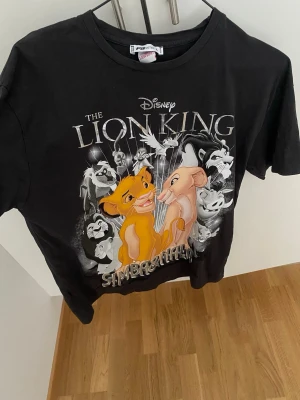 Svart Lion King t-shirt - Säljer en svart t-shirt med Disney's The Lion King-tryck framtill. Motivet visar Simba och Nala i färg med flera andra karaktärer i bakgrunden. T-shirten har rund hals och korta ärmar, perfekt för dig som älskar klassiska Disneyfilmer. Stl L eller ocersize