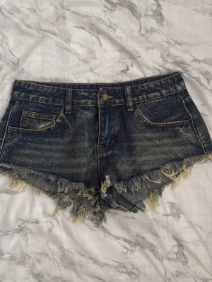 Mörkblå jeansshorts med fransar - Säljer ett par mörkblå jeansshorts med råa, fransiga kanter och slitningar både fram och bak. Shortsen har klassisk femficksdesign, bälteshällor och knappgylf. Perfekta för sommaren och ger en avslappnad vibe. Passar Xs ❤️