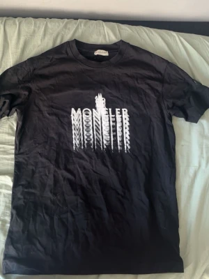 Svart Moncler t-shirt  - Snygg svart t-shirt från Moncler med ett vitt grafiskt tryck på bröstet. Klassisk rund hals och korta ärmar. Perfekt för dig som vill ha en stilren men ändå unik look. Tillverkad i mjuk bomull för skön känsla hela dagen. Kan passa M 