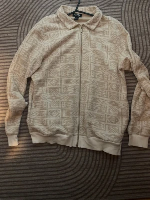 Beige mönstrad ziptröja MAN - Beige ziptröja från MAN med diskret geometriskt mönster över hela plagget. Tröjan har ribbade muddar, krage och dragkedja framtill. Perfekt för lager på lager och en avslappnad stil.