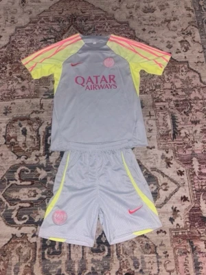 PSG träningsset grå/gul - Säljer ett PSG träningsset med t-shirt och shorts i ljusgrått med neon-gula och rosa detaljer. Tröjan har Qatar Airways-tryck och PSG-logga, samt rosa Nike-märke. Materialet är lätt och andas, perfekt för fotbollsträning eller gym.