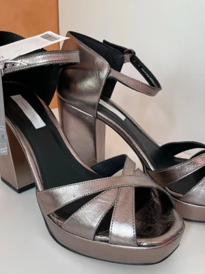 Metallic platåpumps med remmar, äkta skinn, stl 40 - Snygga metallicfärgade platåpumps från H&M med bred klack och öppen tå. Skorna har korslagda remmar framtill och justerbar ankelrem med spänne. Tillverkade i läder för en lyxig känsla och extra hållbarhet. Stl 40, nya oanvända 