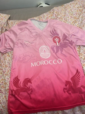 Rosa Morocco sporttröja med enhörningar - Unik rosa sporttröja med Morocco-tryck, Pegasus- och enhörningsmotiv samt molndetaljer. Tröjan har V-ringning och är tillverkad i ett lätt, snabbtorkande material som passar perfekt till träning eller match.