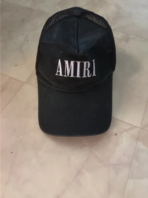 Amiri Keps - Säljer en svart keps från Amiri med vit broderad logga framtill. Kepsen har klassisk böjd skärm och meshmaterial på sidorna och bak för extra ventilation. Perfekt för en streetwear-look.
