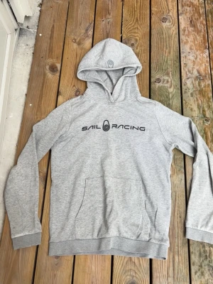 Grå hoodie från Sail Racing - Snygg grå hoodie från Sail Racing med svart logga på bröstet. Klassisk känguruficka framtill och ribbade muddar vid ärmar och nederkant. Tillverkad i mjukt sweatshirtmaterial och har en stor huva för extra chill vibe.