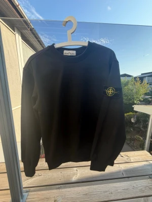 Svart sweatshirt från Stone Island - Svart sweatshirt från Stone Island med klassisk rund hals och ikonisk patch med logga på vänster ärm. Tröjan har ribbade muddar vid ärmslut och nederkant. Perfekt för en stilren och avslappnad look.