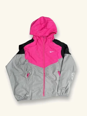 Nike vindjacka rosa och grå - Snygg vindjacka från Nike i färgerna rosa, grå och svart. Jackan har huva, dragkedja framtill och två fickor med dragkedja. Ikonisk Nike-logga på bröstet och elastiska muddar vid ärmslut. Perfekt för en sportig och trendig look.