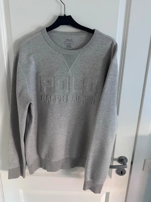 Grå sweatshirt från Polo Ralph Lauren - Snygg grå sweatshirt från Polo Ralph Lauren med broderad logga framtill. Klassisk rund halsringning och ribbade muddar vid ärmslut och nederkant. Perfekt för en avslappnad och stilren look. Använd fåtal gånger och storlek M. Tveka inte att höra av dig för fler frågor!