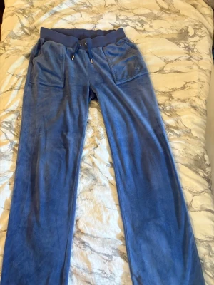 Blå Jucy  mjukisbyxor med fickor - Blå mjukisbyxor i mjuk velour med bred resår i midjan och dragsko. Två stora fickor fram och två bakfickor med knapp. Perfekta för chill dagar eller när du vill vara extra bekväm.