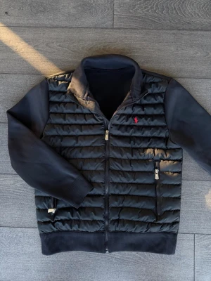 Marinblå Ralph Lauren cardigan  - Snygg svart cardigan från Polo Ralph Lauren med quiltad front och stickade ärmar. Jackan har två dragkedjeförsedda fickor, ribbade muddar och en liten röd logga på bröstet. Perfekt för kalla vinterdagar och riktigt stilren look.